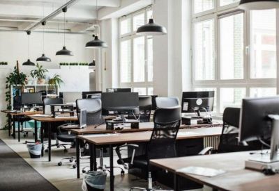 Sunlit open-plan office with rows of wooden desks and black ergonomic chairs.