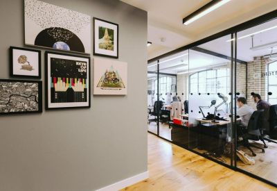 Hallway view of a gallery wall with framed art leading into a glass-partitioned workspace.