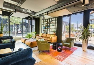 Bright coworking lounge with leather sofas, green armchairs, and floor-to-ceiling windows.