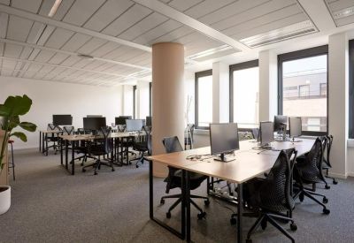 Bright open-plan office space with rows of wooden desks, ergonomic black chairs, and large windows.