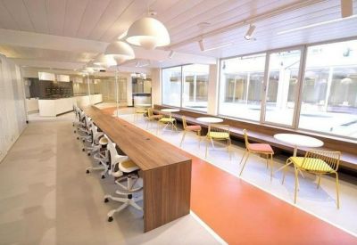 Open-plan coworking area with a long wooden desk, white office chairs, and orange floor accents.