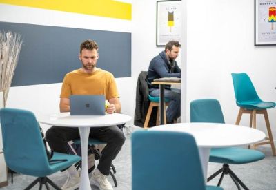 Two people working on laptops at small round tables in a modern work lounge.