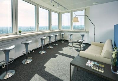 Sunlit breakroom with bar stools, a white sofa, and panoramic city views through large windows.