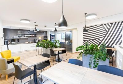 Bright communal lounge and kitchen with yellow and grey armchairs, plants, and modern lighting.