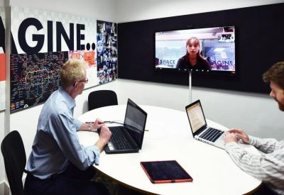 Meeting room with two people using laptops and a screen displaying a video conference call.