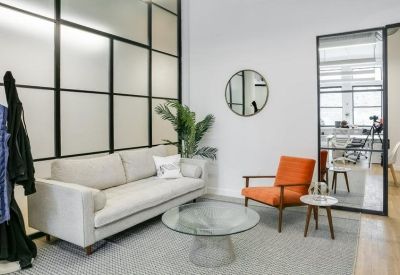 Modern reception lounge with a light grey sofa, orange armchair, and glass-partitioned walls.