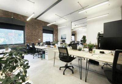 Sunlit open-plan office with white desks, ergonomic chairs, and large industrial windows.