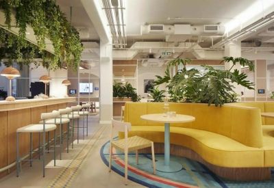 Bright communal lounge featuring a curved yellow sofa, white cafe tables, and lush indoor plants.