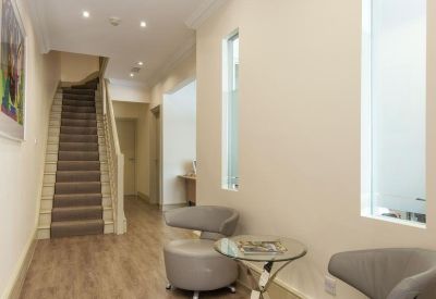 Bright reception hallway with modern grey armchairs, wooden flooring, and a staircase.