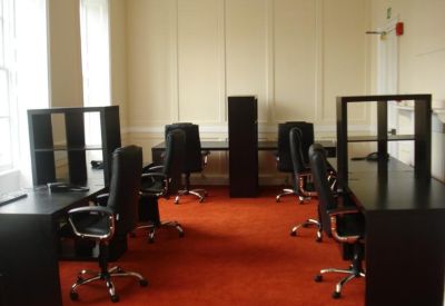 Brightly lit office room featuring dark desks, black ergonomic chairs, and red carpeting.