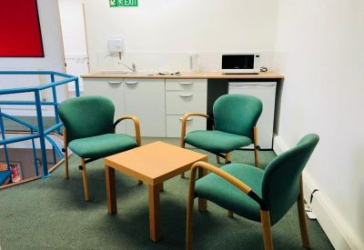 Communal breakout area with green armchairs, a wooden table, and a small kitchen unit.