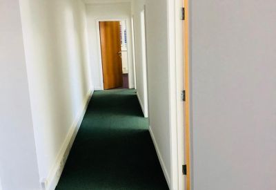 Long office corridor with white walls and green carpeting leading to a wooden door.