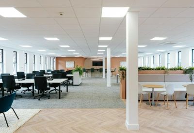 Open-plan office space featuring white desks, black chairs, and a breakout area with plants.