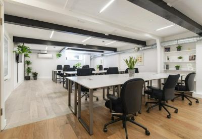 Open-plan office space with long white desks, black ergonomic chairs, and indoor plants.