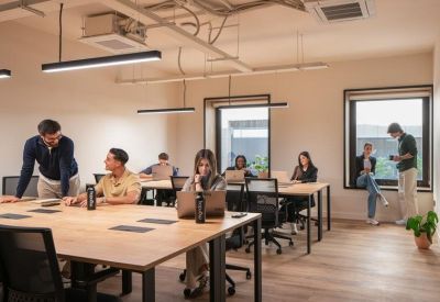 Coworking space with long wooden desks, ergonomic chairs, and people working near large windows.