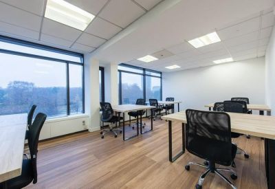 Modern open-plan office with light wood desks, black ergonomic chairs, and large windows.