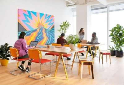 Spacious open-plan coworking area with long wooden tables, vibrant art, and potted plants.