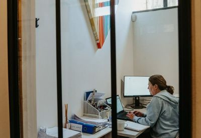 Private glass-fronted office featuring a person working at a desk with dual monitors.