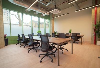 Sun-drenched open-plan workspace with long wooden desks and ergonomic black mesh chairs.