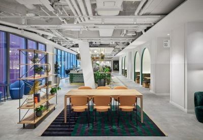 Open-plan coworking space featuring a long wooden table, orange chairs, and exposed ceiling pipes.