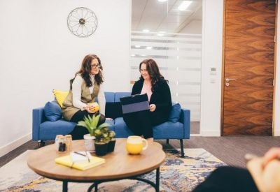 Two women talking on a blue sofa in a lounge area with a wooden coffee table.
