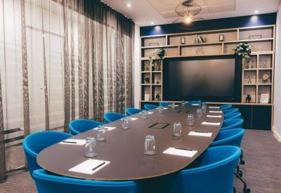 Oval boardroom table with blue chairs and a large wall-mounted screen.