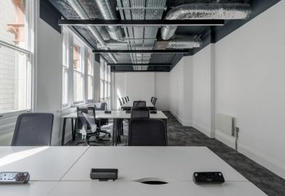 Open-plan office space with white desks, black ergonomic chairs, and exposed ceiling pipes.