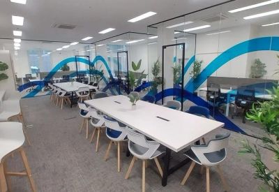 Spacious communal coworking area with long white tables, blue wave wall graphics, and potted plants.