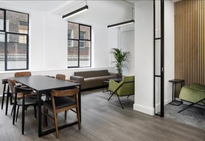 Breakout space with a long dining table, grey sofa, and green armchairs near large windows.