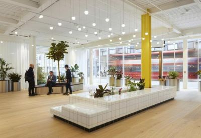 Spacious communal lounge with light wood floors, potted trees, and rows of glowing pendant lights.