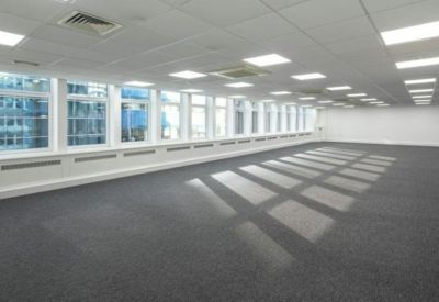 Spacious, empty open-plan office with grey carpeting and large windows providing natural light.