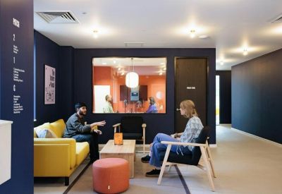 Casual meeting area with a yellow sofa and navy blue walls looking into a glass-walled studio.