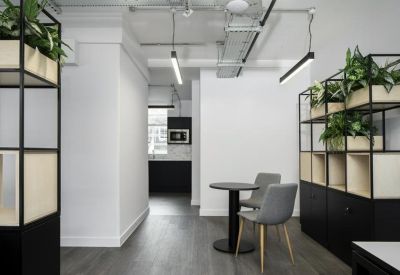 Bright breakout space with a round table, grey armchairs, and lush green plants on shelving.