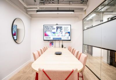 Bright meeting room with a long table, pink chairs, and a wall-mounted screen.