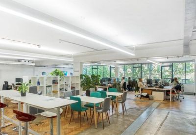 Open-plan coworking space with long white tables, teal chairs, and wooden parquet flooring.