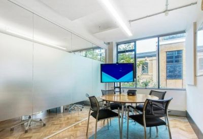 Sunlit meeting room with a circular wooden table, black chairs, and a large wall-mounted screen.