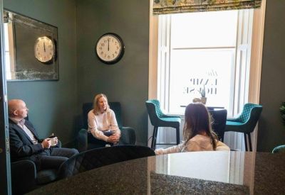 Breakout lounge area with teal velvet chairs, a wall clock, and people chatting by a large window.