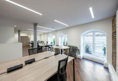 Spacious coworking area with large arched windows, long wooden tables, and bright white walls.