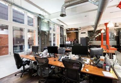 Bright open-plan office with wooden desks, black ergonomic chairs, and large windows.