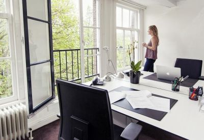 Bright office space with white desks and large windows overlooking lush green trees.