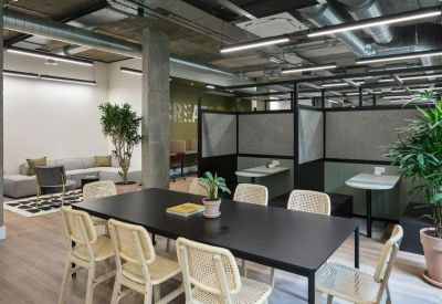 Bright office space featuring private workstations, a common table with woven chairs, and indoor plants.