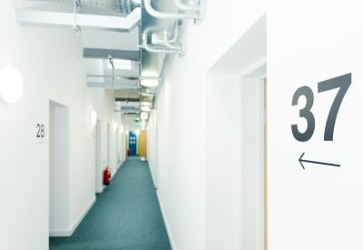 Bright white corridor with exposed industrial ceiling pipes and a blue carpeted floor.