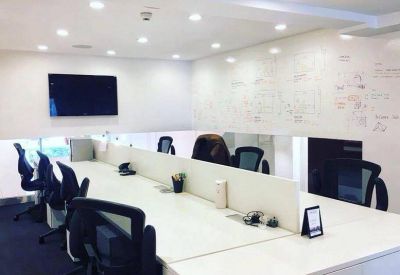 Open-plan office space with white shared desks, black ergonomic chairs, and whiteboard walls.