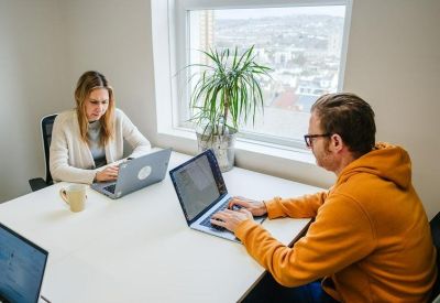 Bright shared workspace with two people working at white desks by a large window.