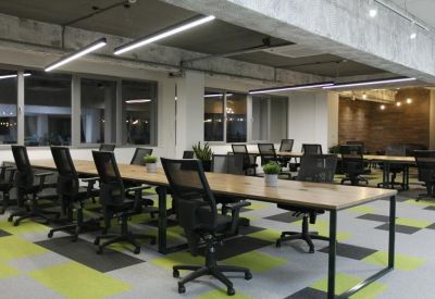 Bright open-plan workspace with long wooden desks, ergonomic black chairs, and colorful geometric carpet tiles.
