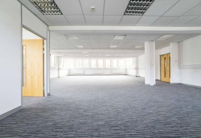 Spacious open-plan office with grey carpets, suspended ceilings, and rows of windows.