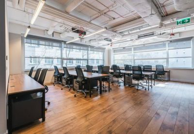 Bright open-plan office with rows of wooden desks, black chairs, and large windows.