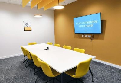 Modern meeting room with a large white table, yellow chairs, and a wall-mounted TV screen.