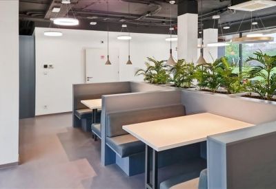 Modern communal breakout area with grey booth seating, light wood tables, and indoor plants.