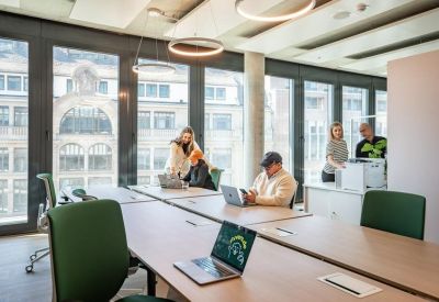 Bright open-plan workspace with long wooden desks and city views through large windows.